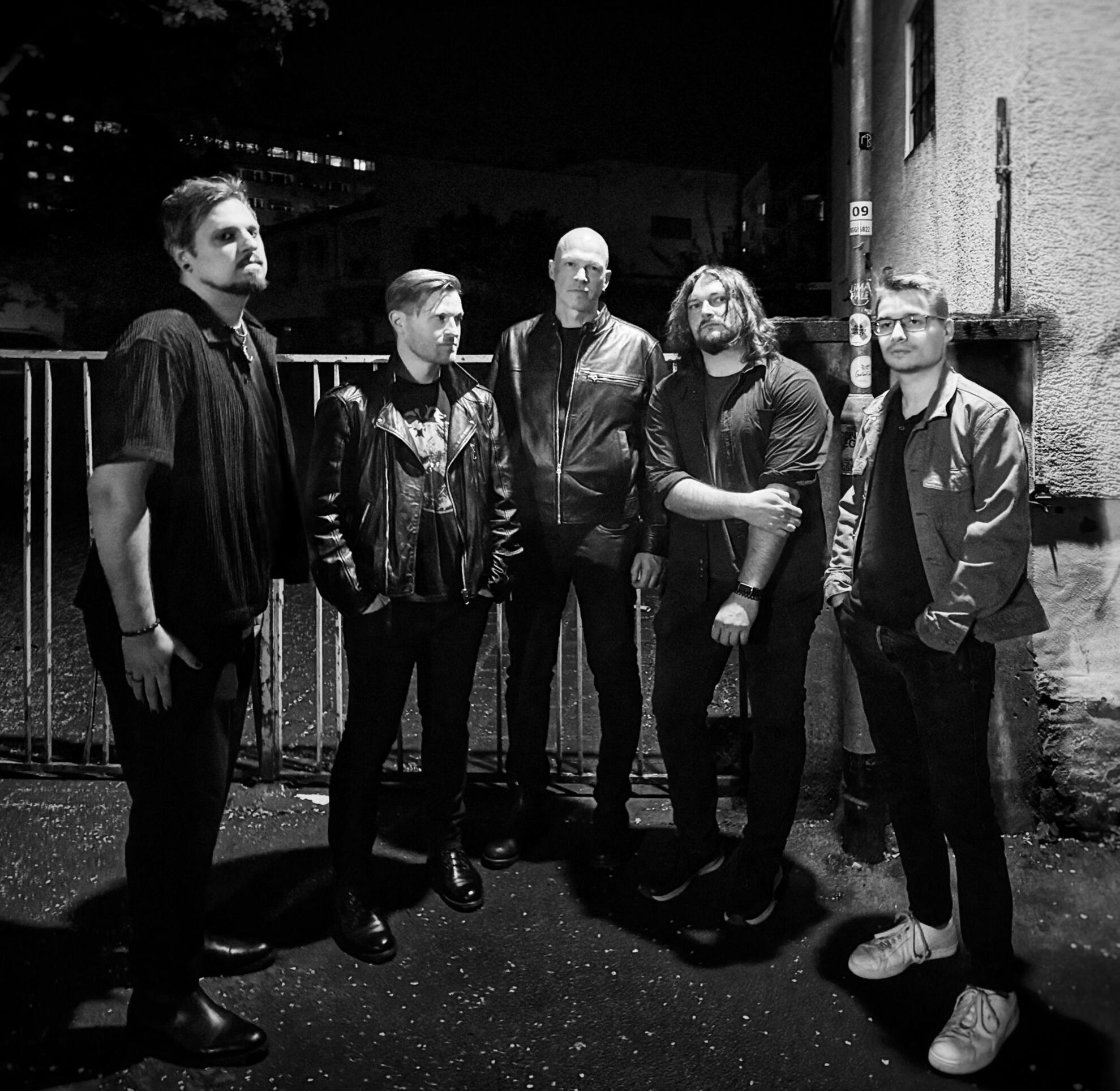Our Visit To The Moon band photo April 2026 Black and white photo of the five male bandmembers of Our Visit To The Moon in dark clothing posing in front of a church wall